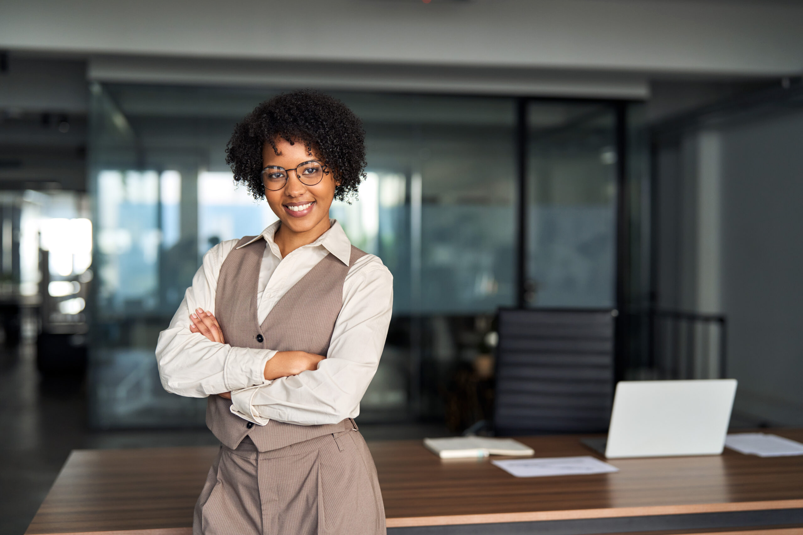 Confident happy young African business woman standing in office, portrait. Confident smiling young professional African American business woman, happy businesswoman corporate leader, female lawyer or hr manager standing arms crossed in office looking at camera, portrait.