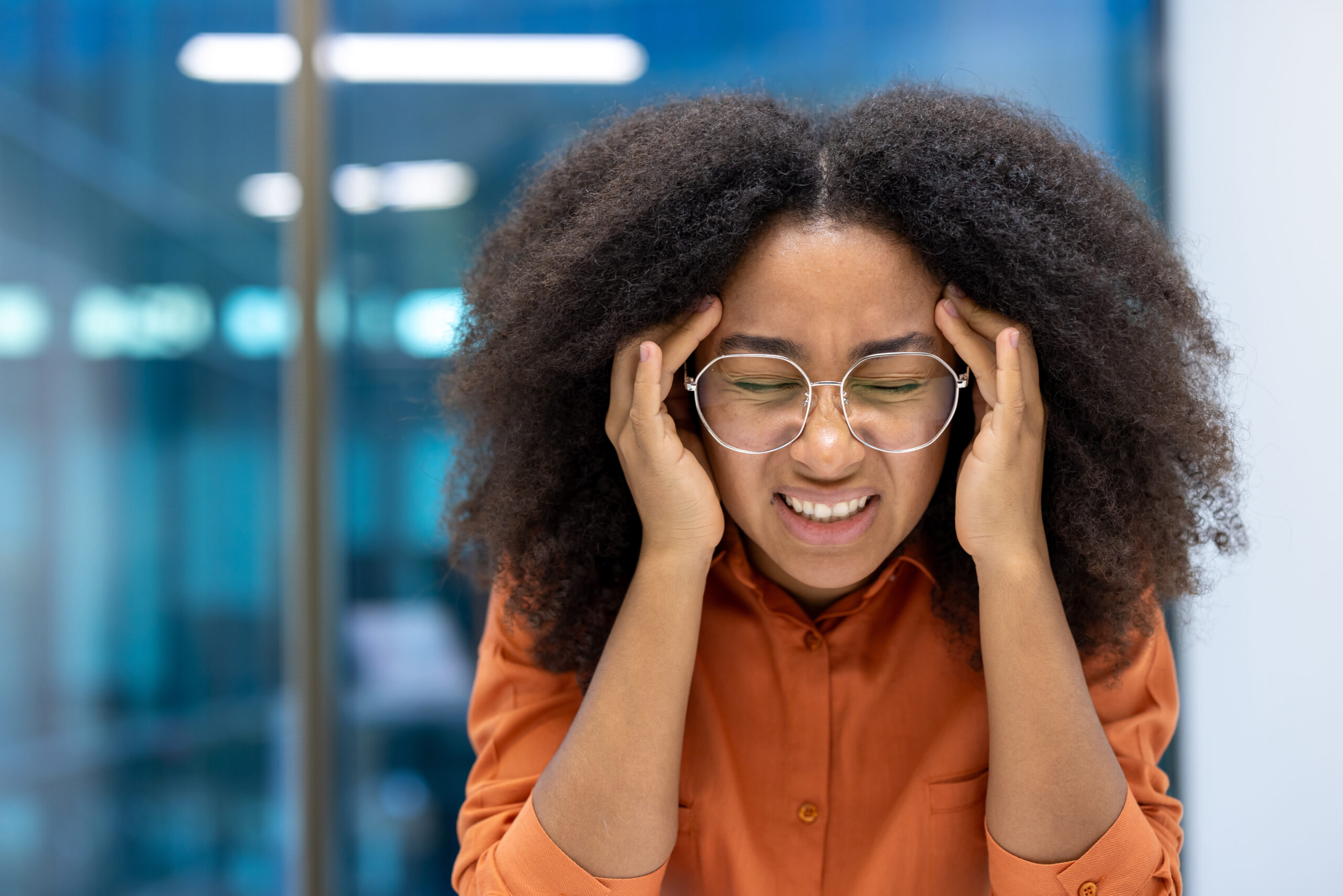 Headache, sick overworked and stressed woman at workplace close up. Businesswoman holding her head with hands while sitting at desk inside office